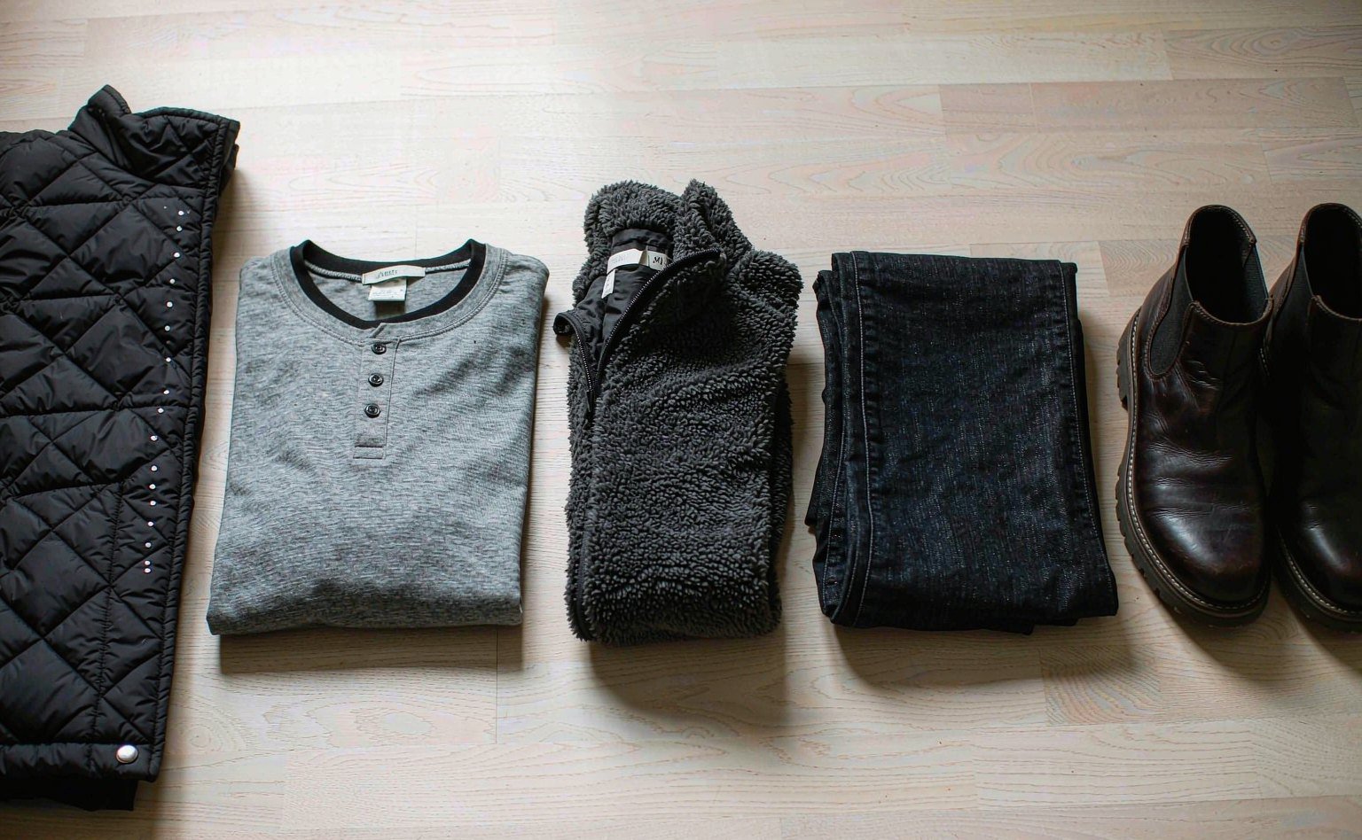 man's winter outfit laid flat: black puffer jacket, grey merino base layer, charcoal fleece mid layer, on pale oak floor
