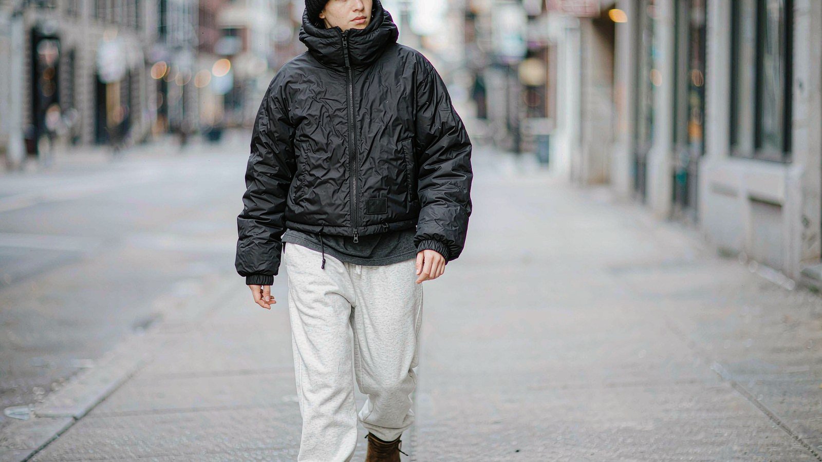 Model in a short black down bomber jacket with a fixed hood standing on a grey city street in cold daylight
