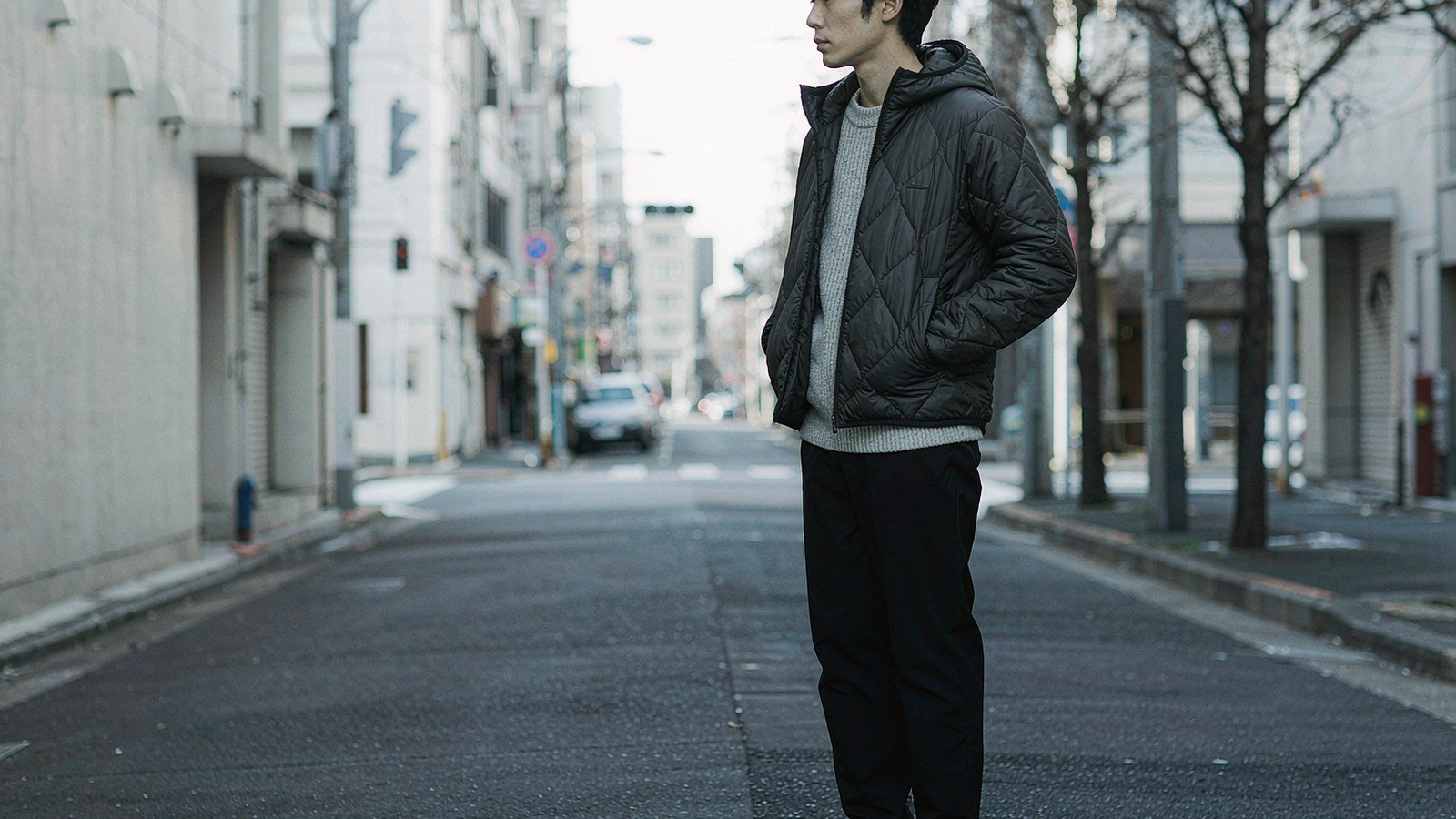 A person wearing a black quilted down hooded jacket on a cold city street, shot from the side.