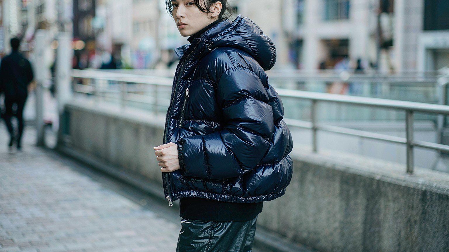 Man wearing a glossy navy short puffer jacket with horizontal quilting on a grey city street in cold daylight