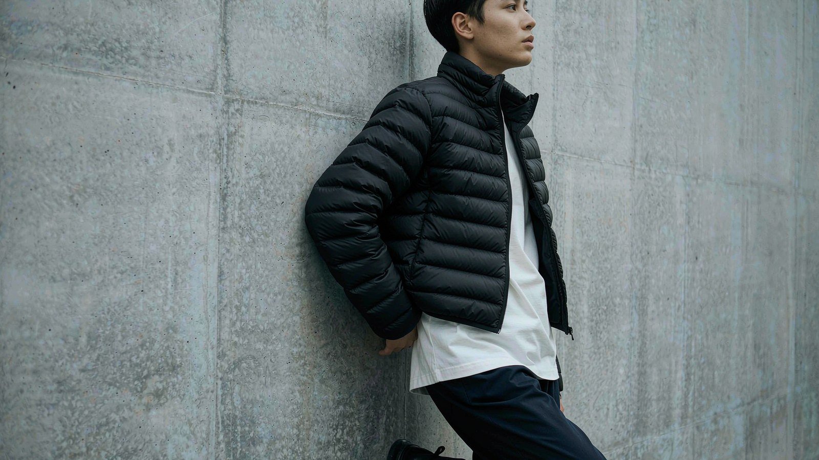 Model wearing a boxy black baffled down puffer jacket against a plain concrete wall