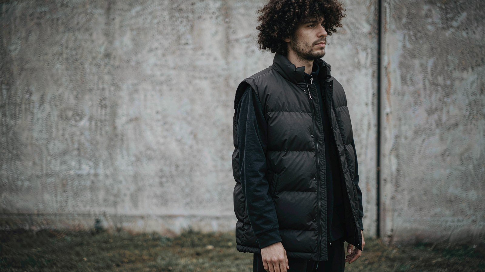 A person in a black matte down vest with a measuring tape across the chest, standing outdoors in cold light.
