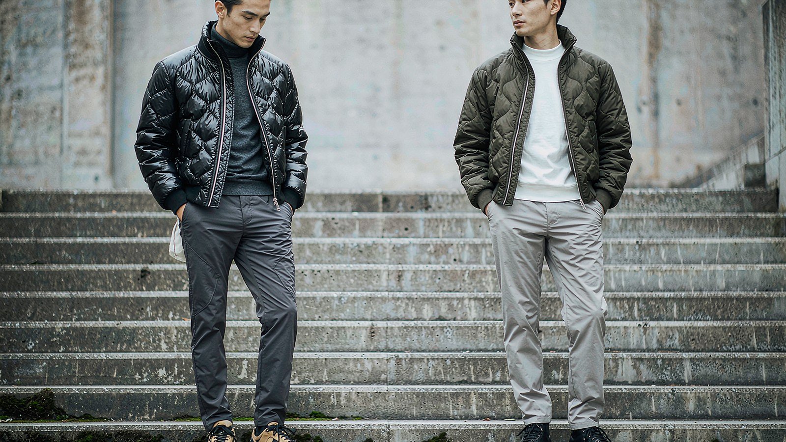 Two models in short quilted bomber jackets standing on a concrete stairway in winter light